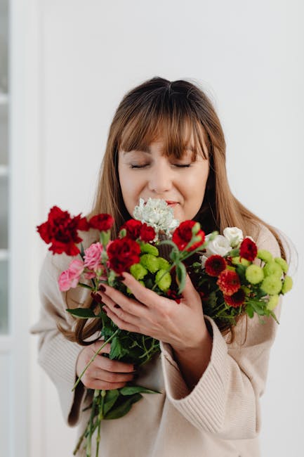A woman with light brown hair and bangs, dressed in a beige cardigan, holds a vibrant bouquet of fresh flowers close to her face with both hands, gently smelling the blooms. The bouquet features a mix