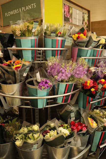 A display of mixed floral bouquets arranged in metal and striped paper-wrapped buckets situated on a tiered metal stand inside a florist shop. The bouquets feature a variety of fresh flowers, includin