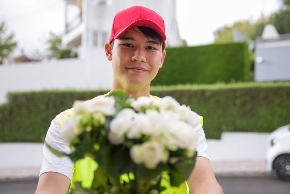 A delivery person wearing a red cap and a white T-shirt holding a vibrant bouquet of fresh white roses and greenery, with some flowers partially wrapped in clear cellophane, ready for flower delivery.