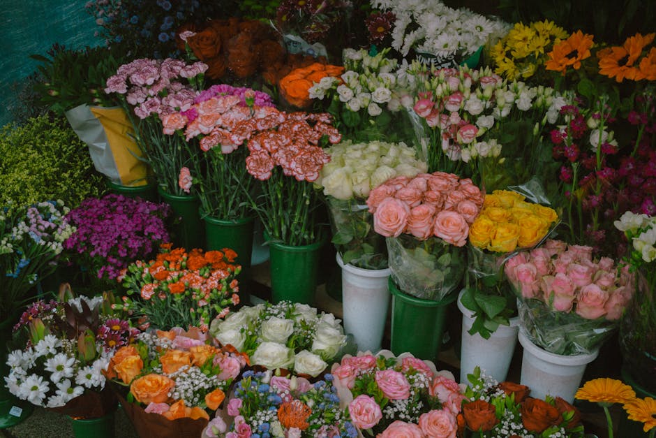 A vibrant display of fresh flowers arranged in a variety of bouquets and potted plants, including pink, white, yellow, orange, and purple blossoms. The bouquets feature roses, carnations, daisies, and
