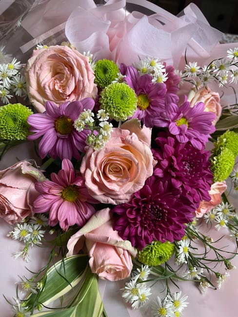 A vibrant floral arrangement featuring light pink roses with tightly curled petals, purple daisies with bright yellow centers, small white daisy-like flowers with yellow centers, and vivid green chrys