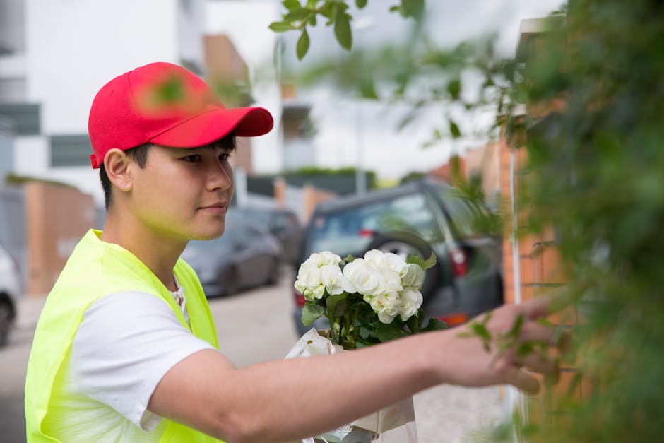 A young delivery person wearing a red cap, a high-visibility neon yellow vest over a white t-shirt, is holding a bouquet of fresh white hydrangeas and roses wrapped in transparent cellophane. They are