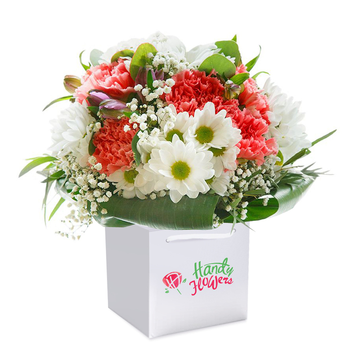 A vibrant floral bouquet, evocative of the best of Sipson's local charm, stands elegantly upright in a clean, square white gift bag, its freshness and vitality captured against a spotless background. At the heart of the arrangement are coral-pink and fiery red carnations, their distinctive ruffled petals layered for lush, tactile appeal, reminiscent of colourful borders seen along Sipson Lane. Scattered between, pure white daisies with striking lime-yellow centers provide a cheerful lift, their neat outlines echoing the tidy hedgerows of nearby Harmondsworth. Cloud-like clusters of baby's breath intermingle throughout, softening the composition and imparting a floating, whimsical quality. On the bouquet's left, partially formed buds dusted with purple hues suggest new beginnings and the turning of seasons. Abundant deep green foliage fans elegantly at the base, the broad, glossy leaves gently curving to gather all elements together in harmonious balance. The bouquet is contained within a stylish white gift bag, finished with sturdy white handles-perfect for carrying along Sipson Road to a friend's celebration or delivering a smile on a quiet afternoon. Every stem is precisely placed, highlighting the skilled hand of a local florist and offering a gesture of beauty for birthdays, significant milestones, or thoughtful surprises across Sipson. The overall effect combines premium floristry with the authentic warmth of the neighborhood.