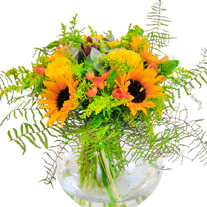 With a palette drawn from the brightest days along Sipson's parkways, this bouquet is both modern and abundant. Large sunflowers dominate the arrangement, their golden petals unfurling around rich, chocolate-brown centers like small suns over Harmondsworth Lane. Interlaced are buttery yellow garden roses, their intricate, swirling petals lending a classic grace to the untamed design. Clusters of shiny red hypericum berries nestle amongst the greenery, tiny beacons of festivity against a tapestry of green and gold. Feathery, arching ferns and slender spikes spill from the bouquet's outline, enhanced by sprigs of goldenrod just beginning to bloom with tiny, vivid yellow blossoms. Bluish eucalyptus leaves, smooth and aromatic, provide contrast and a cool finish. All elements are gathered in a broad, clear glass vase-its transparent round form revealing stems knotted and settling in clear water, adding architectural drama and freshness. The impression is of a bouquet bursting with morning cheer, ideal as a joyful gift to mark new beginnings, anniversaries, or welcome a neighbor to Sipson Village. Every detail, from petal to stem, is a nod to local blooms and the enthusiasm of flower-lovers throughout our Heathrow community.